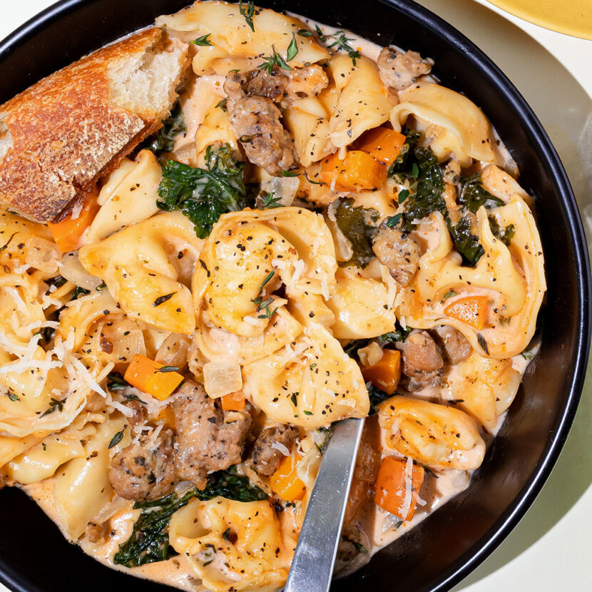 An image of prepared Creamy Sausage & Tortellini Soup made with Italian pork sausage, onion, carrots, garlic, Italian seasoning, Pacific Foods® Organic Free Range Chicken Broth, cream, tomato paste, cheese tortellini and kale.