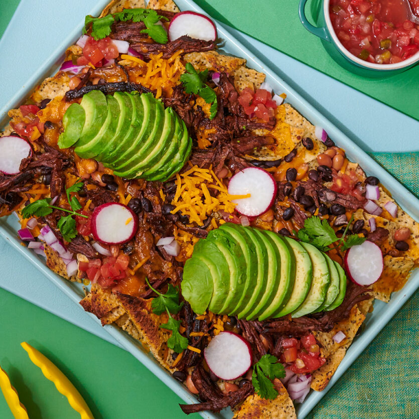 An image of prepared Instant Pot® Shredded Beef Nachos made with Pacific Foods® Beef Broth, chuck roast, tortilla chips, red onion, Cheddar cheese, pico de gallo, black beans, pinto beans, cilantro, radish and avocado.