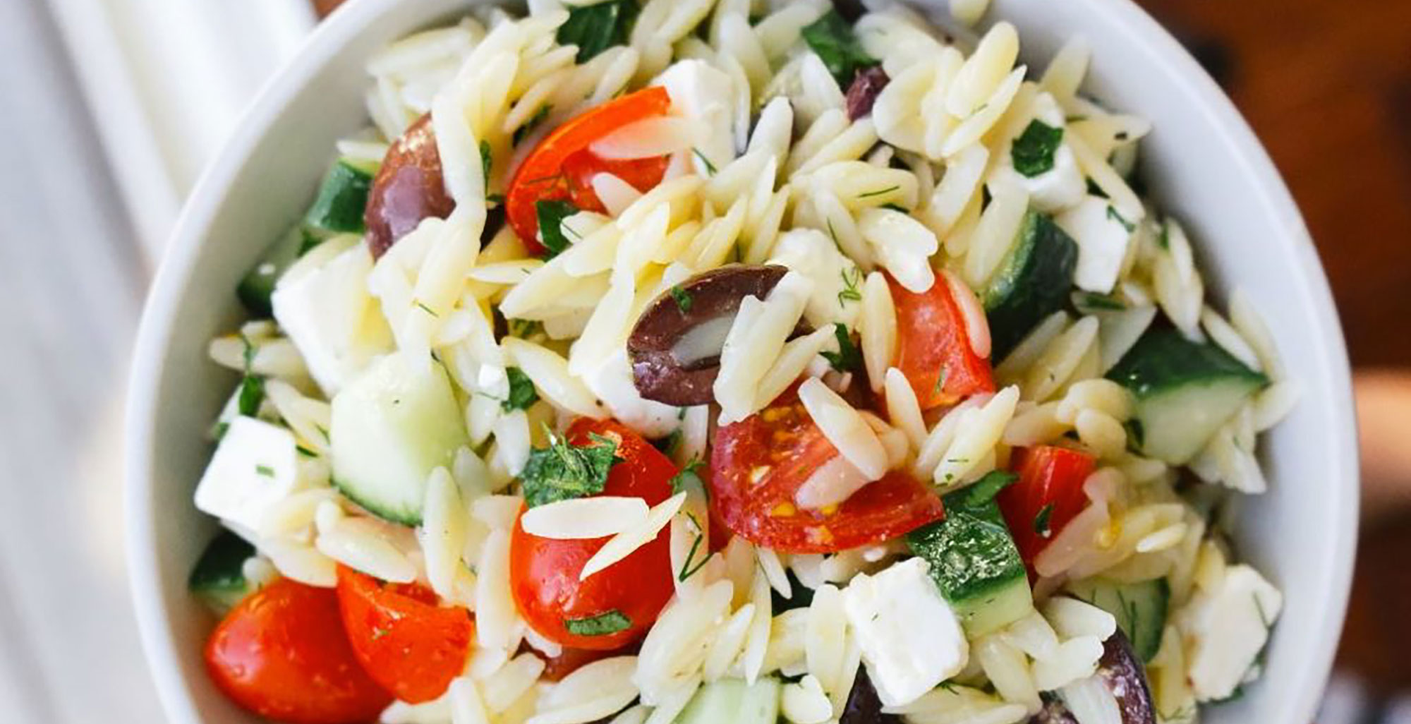 An image of prepared Mediterranean Orzo Salad made with Pacific Foods® Organic Low Sodium Vegetable Broth, orzo pasta, feta cheese, cherry tomatoes, cucumber, kalamata olives, parsley and dill.