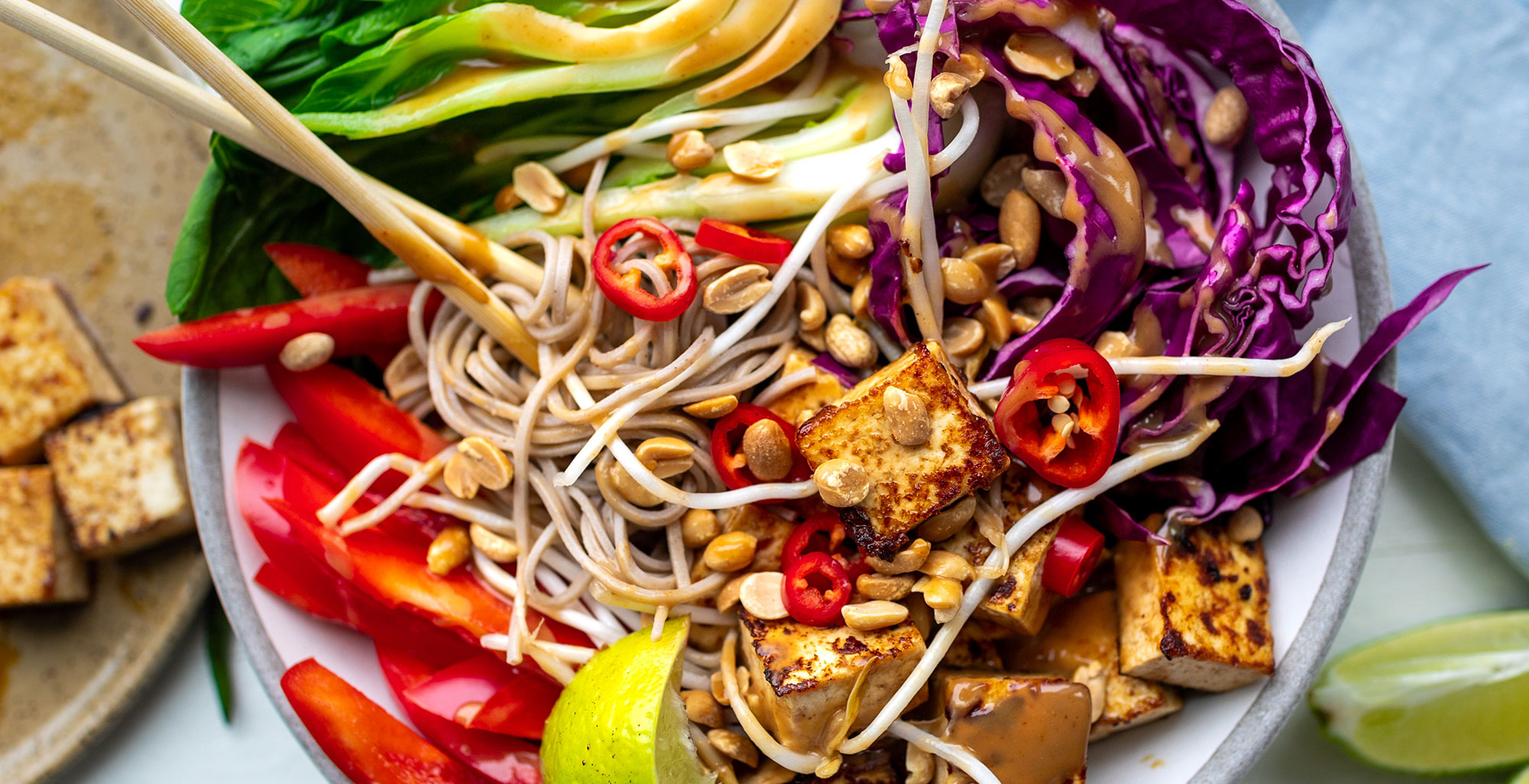 An image of prepared Peanut Tofu Noodle Bowl made with tofu, honey, peanut butter, sesame oil, tamari, Pacific Foods® Organic Mushroom Broth, Japanese soba noodles, bok choy, bean sprouts, red bell pepper, red cabbage, red chili pepper, peanuts and limes.