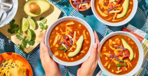 An image of prepared Chicken Enchilada Soup made with Pacific Foods® Organic Condensed Tomato Soup, Pacific Foods® Organic Low Sodium Free Range Chicken Broth, corn, green chiles, chicken, tortilla strips, Cheddar cheese, avocado and cilantro.